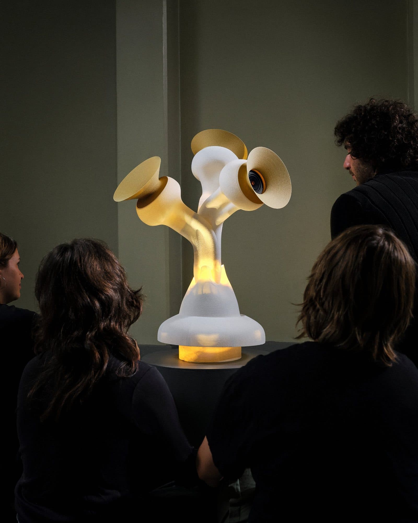 Four people sitting in a dim room around the Entity, a glowing white sculpture with three golden gramophone-like speakers.