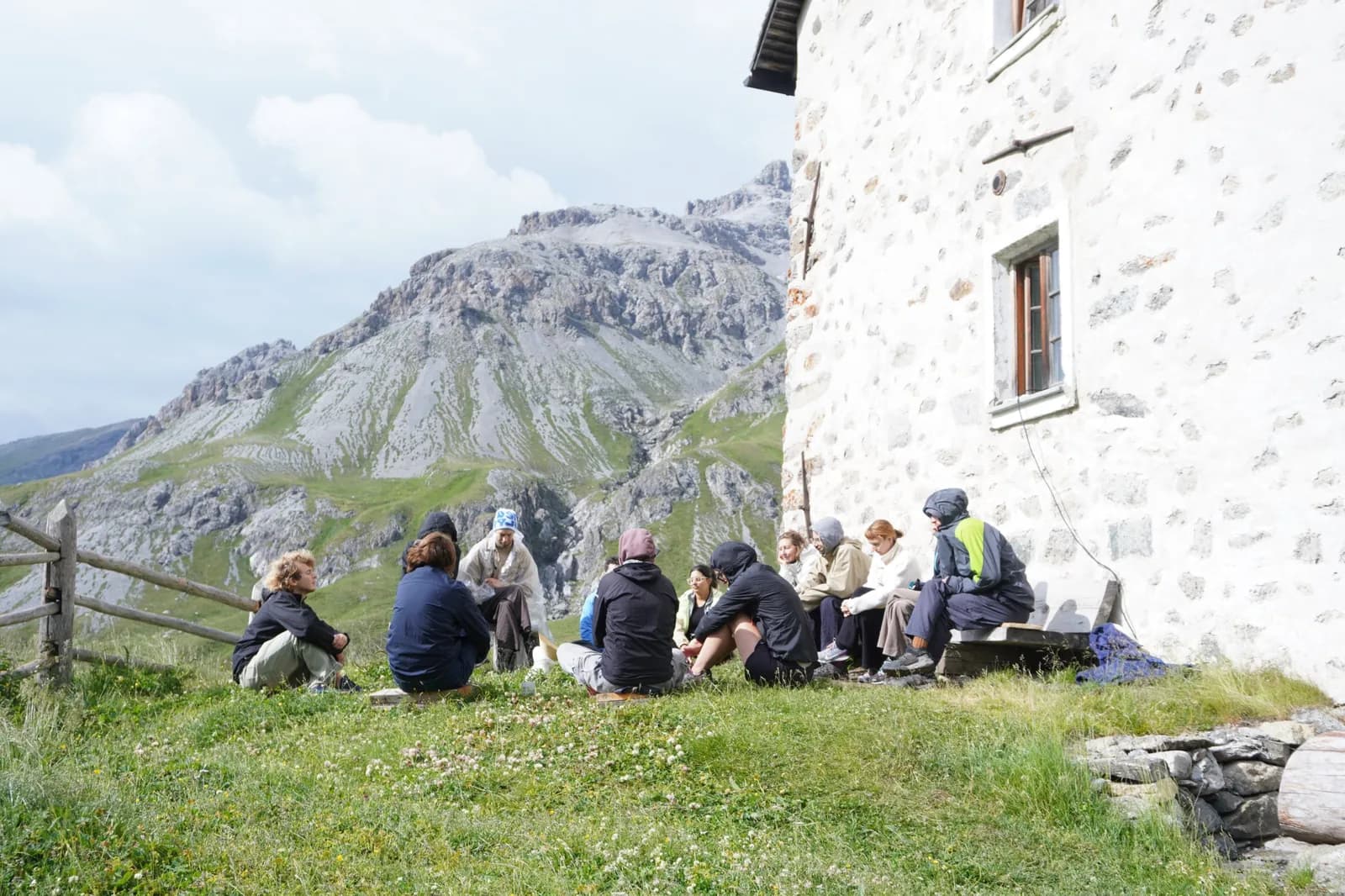 A circle of around fifteen people beside a stone mountain hut, with a rocky peak behind them.