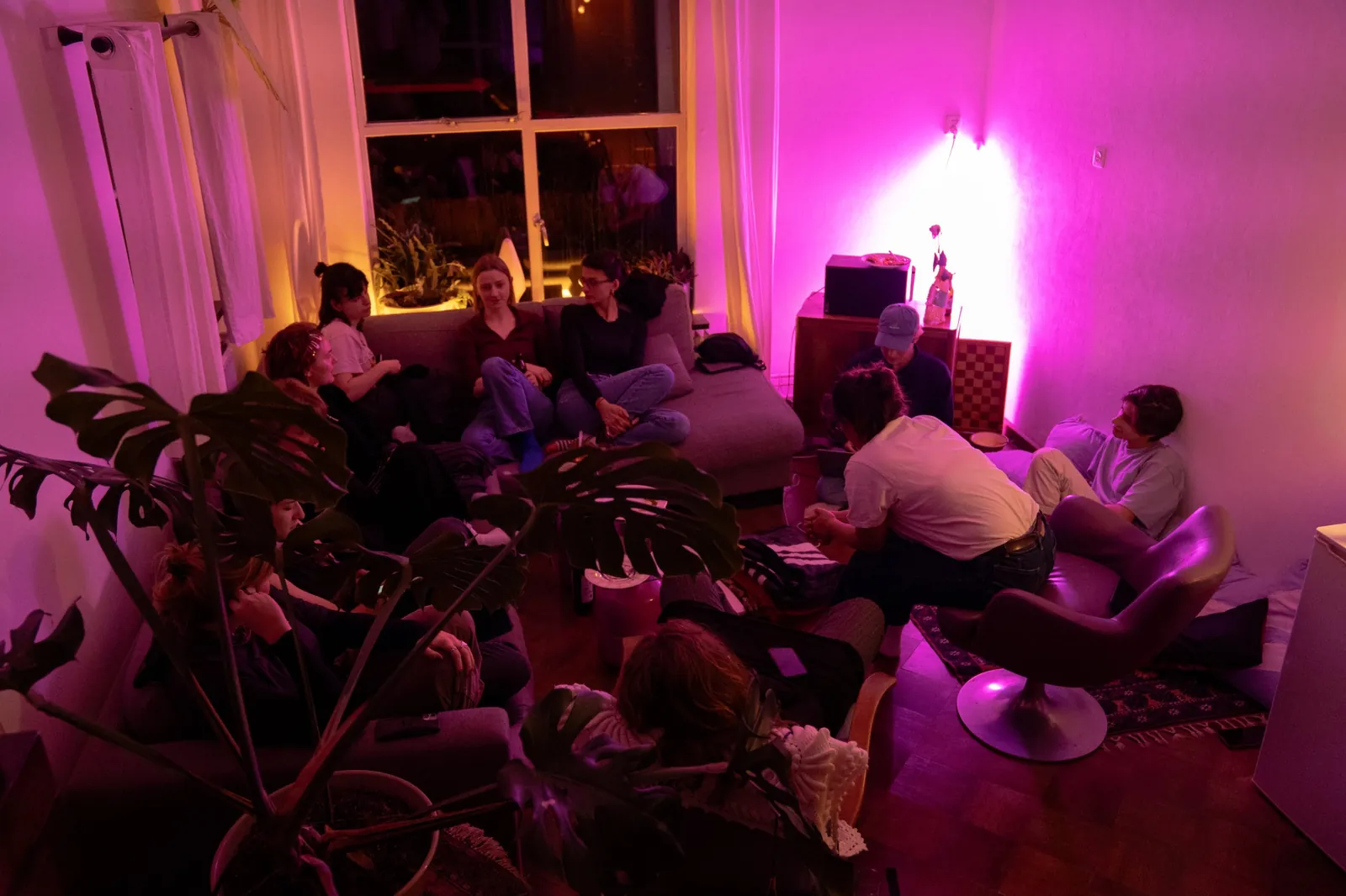 A living room in pink and orange light, a dozen people sitting on sofas, chairs and the floor, listening.