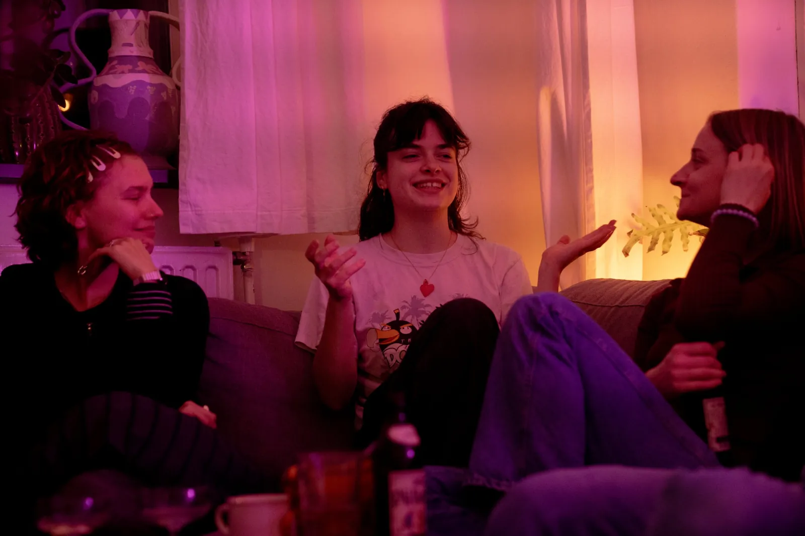 Three people on a sofa in pink light, one of them talking with her hands, the others smiling.